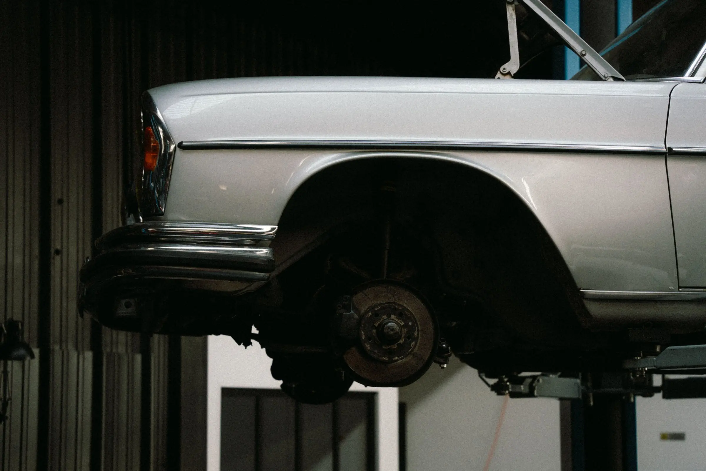 Classic silver sedan raised on a lift in a dim garage bay, front wheel off, brake hub exposed