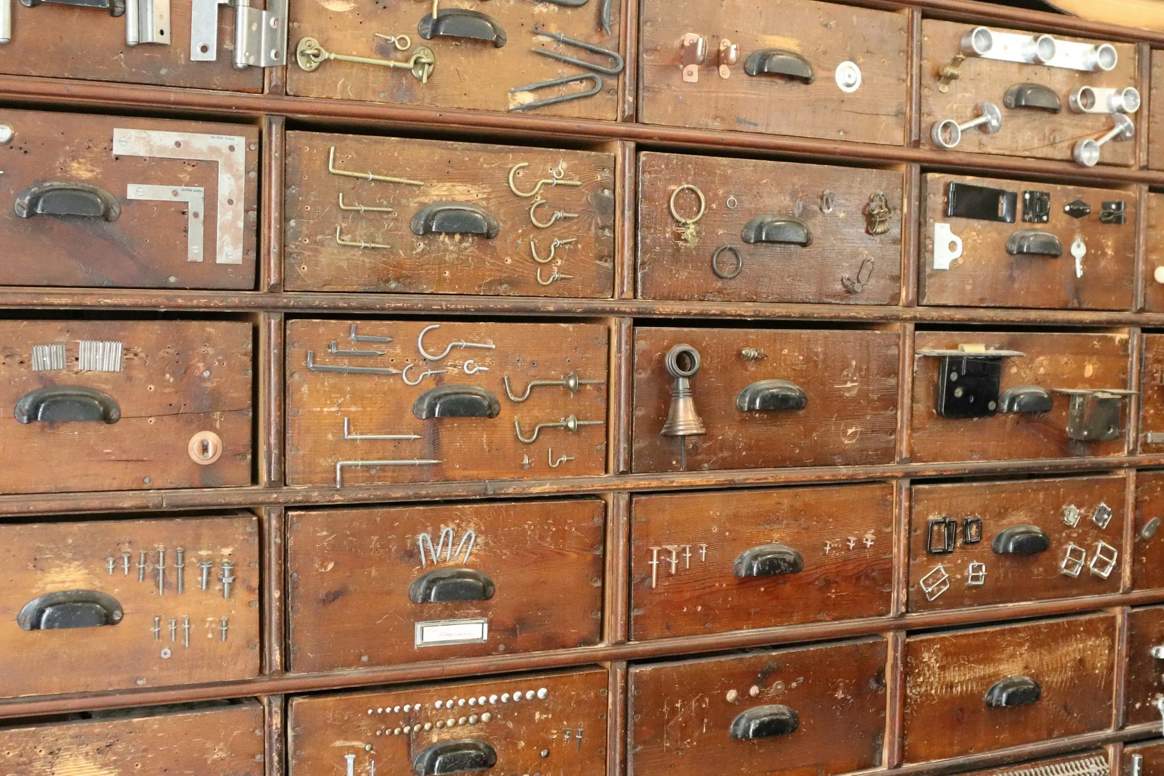 Wall of antique wooden hardware drawers with sample hooks, hinges and latches mounted on the fronts